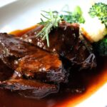 Brasato al vino rosso servito su un piatto, con contorni di verdure e un bicchiere di vino rosso accanto.